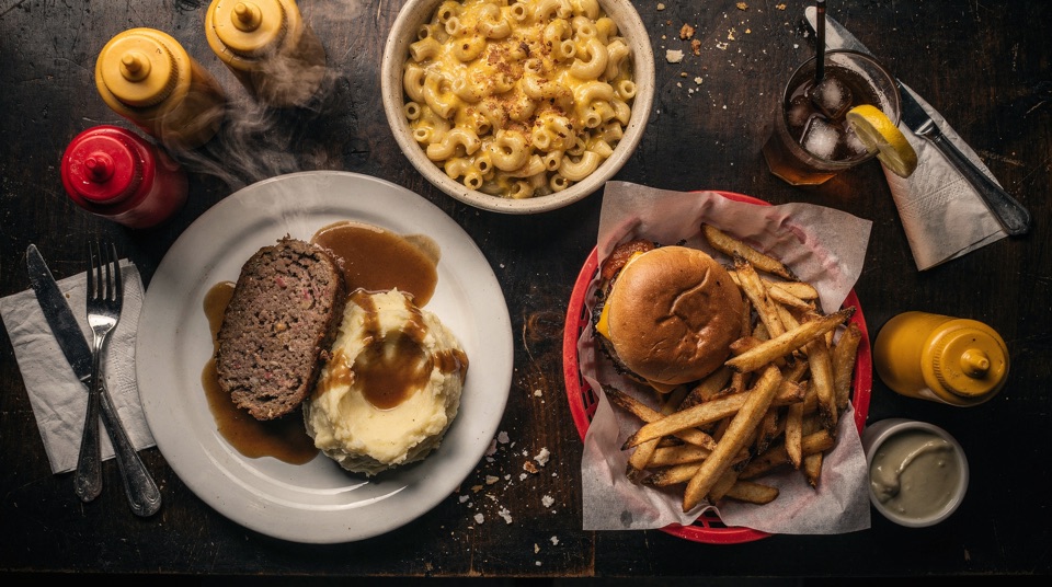 Plates of meatloaf, a burger with hand-cut fries, and mac and cheese on a dark wood table.